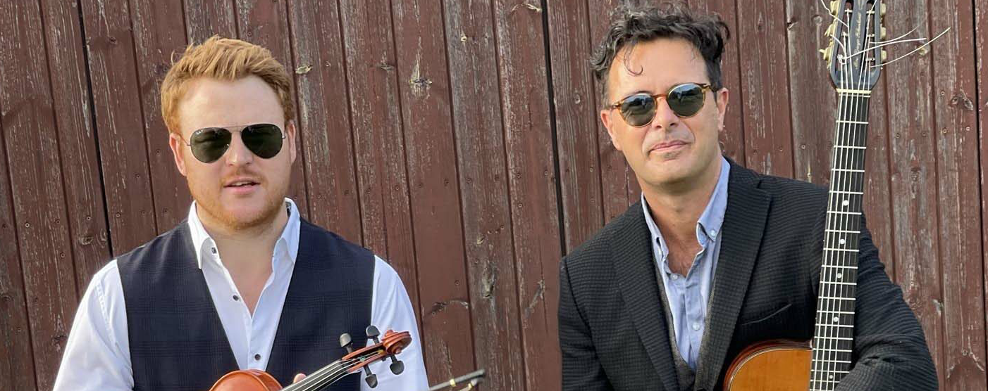 Violinist Ben Holder and guitarist Stuart Carter-Smith standing with their instruments in front of a wooden fence.